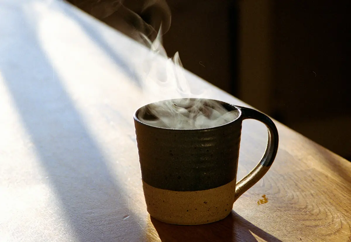 A steaming ceramic mug of coffee on a wooden counter in low morning sun